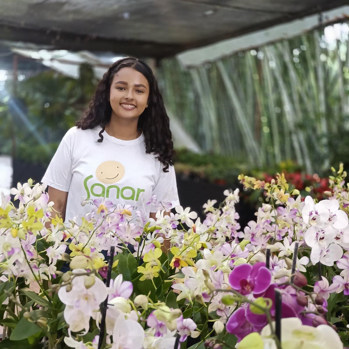 Person standing among orchids in a greenhouse wearing a white shirt with a logo.
